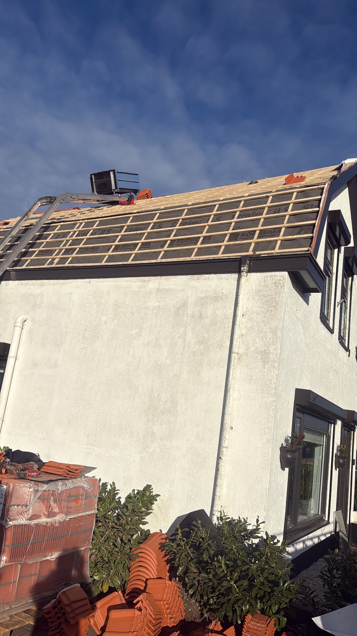 Dach w trakcie krycia dachówką w Budel. Widoczna konstrukcja dachu z łatami i membraną. Na dole stosy dachówek. Prace dekarskie w toku, jasne niebo w tle.