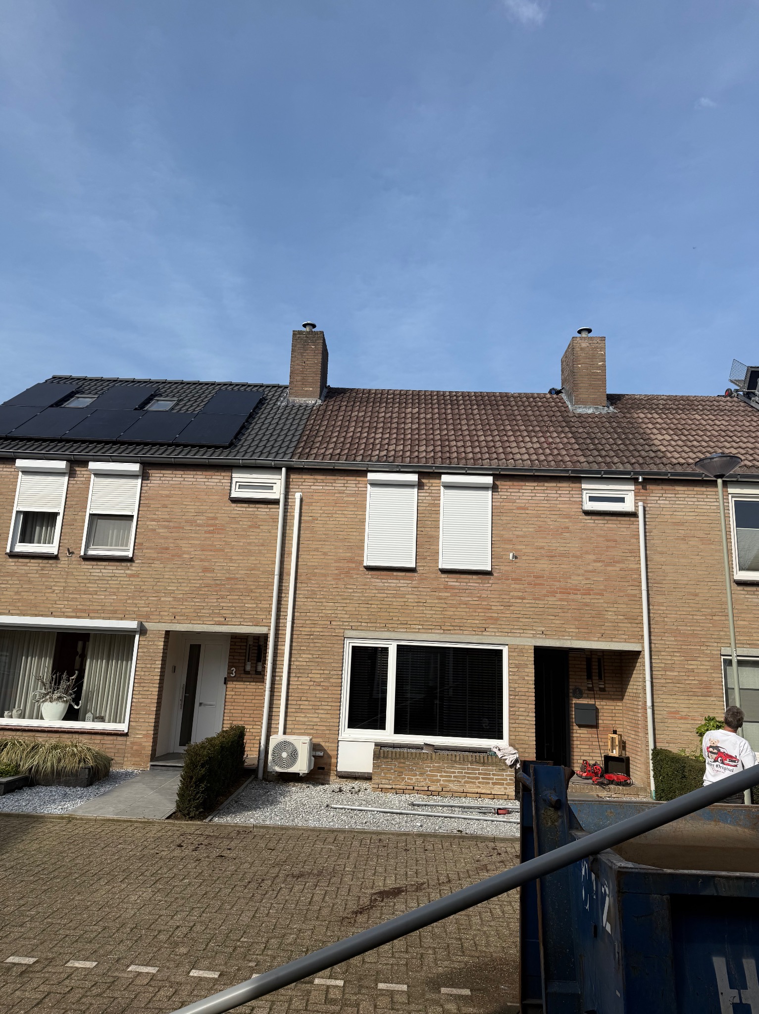 Bliźniak z cegły klinkierowej: część dachu pokryta panelami solarnymi, część dachówką. Widoczny komin i pracownik na dachu. Kontener na gruz na pierwszym planie.