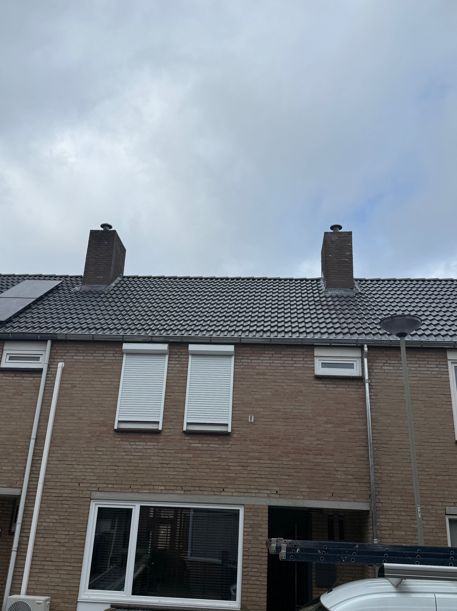 Szary dach z dachówki ceramicznej na dwukondygnacyjnym domu z cegły, z dwoma kominami i roletami w oknach. Widok z dołu na pochmurne niebo.