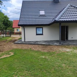 ARIEL LEWANDOWSKI USŁUGI REMONTOWE - Elewacja domu jednorodzinnego z ciemną dachówką i kostką brukową wokół. Widoczne okno dachowe i fragment ogrodu. Dom po remoncie, świeżo pomalowany.
