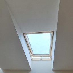 ARIEL LEWANDOWSKI USŁUGI REMONTOWE - Widok z dołu na okno dachowe w jasnym pomieszczeniu. Białe ściany i sufit tworzą minimalistyczne wnętrze. Okno w drewnianej ramie wpuszcza naturalne światło.