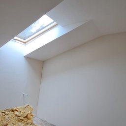 ARIEL LEWANDOWSKI USŁUGI REMONTOWE - Poddasze po gładziowaniu ścian i sufitu, z oknem dachowym i stertą wełny mineralnej w rogu. Ściany pomalowane na jasny kolor, podłoga zabezpieczona.