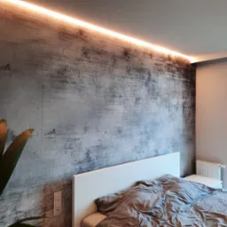 Ściana w sypialni wykończona w stylu betonu architektonicznego z podświetleniem LED, widoczna roślina doniczkowa i fragment łóżka.