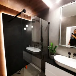 Nowoczesna łazienka z kabiną typu walk-in z czarnymi profilami, umywalkami nablatowymi, podświetleniem LED i elementami imitującymi drewno. W lustrze odbija się osoba robiąca zdjęcie.
