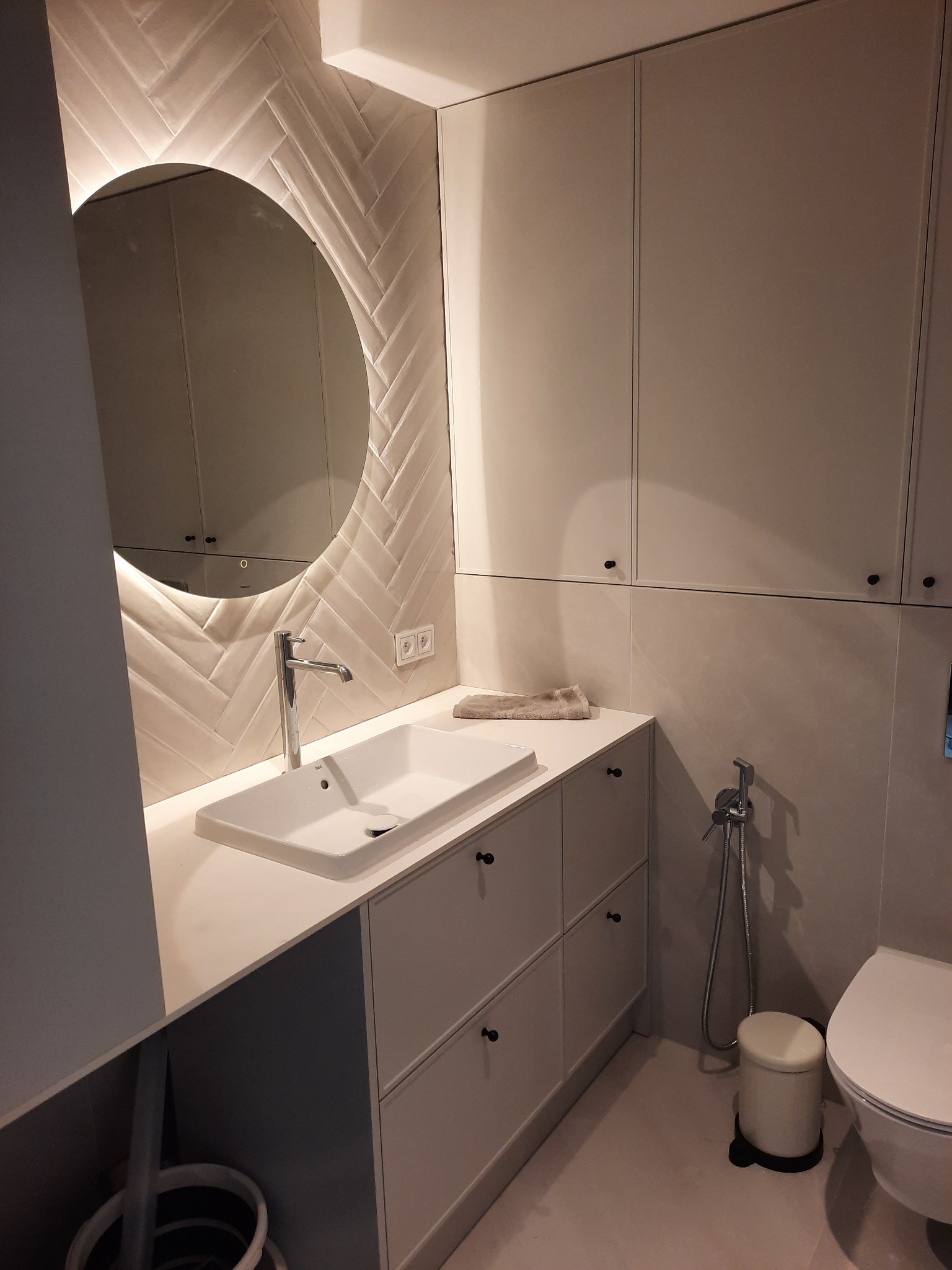 Nowoczesna łazienka z umywalką nablatową, okrągłym podświetlanym lustrem na ścianie wyłożonej płytkami w jodełkę, oraz zabudową meblową w jasnych odcieniach.