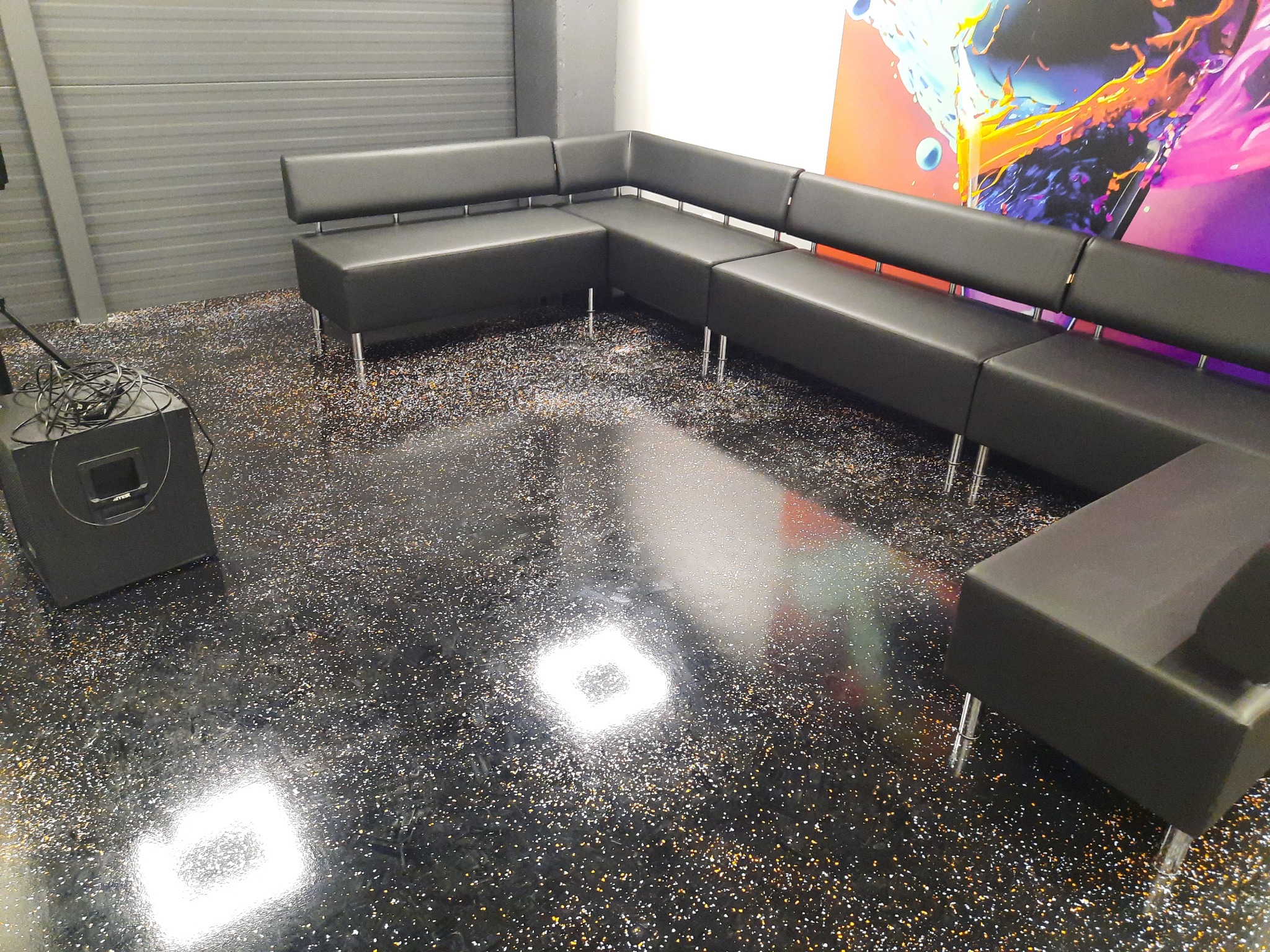 Ciemny, modułowy narożnik z ekoskóry na metalowych nóżkach, ustawiony w pomieszczeniu z błyszczącą, żywiczną posadzką w odcieniach czerni i złota, widoczny głośnik i fragment abstrakcyjnej grafiki...