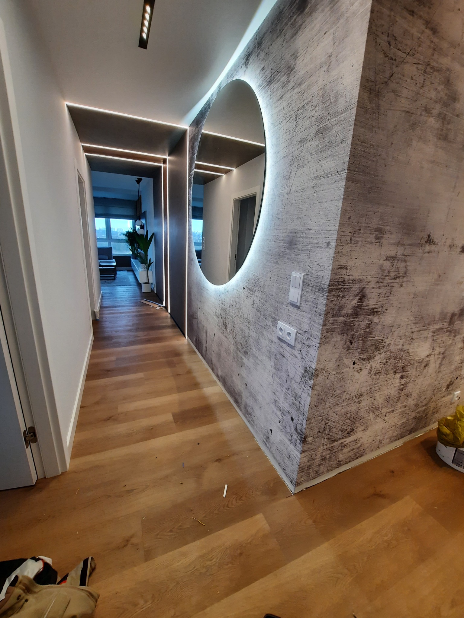 Nowoczesny korytarz z oświetleniem LED, okrągłym lustrem z podświetleniem, ścianą wykończoną imitacją betonu i parkietem z jasnego drewna.