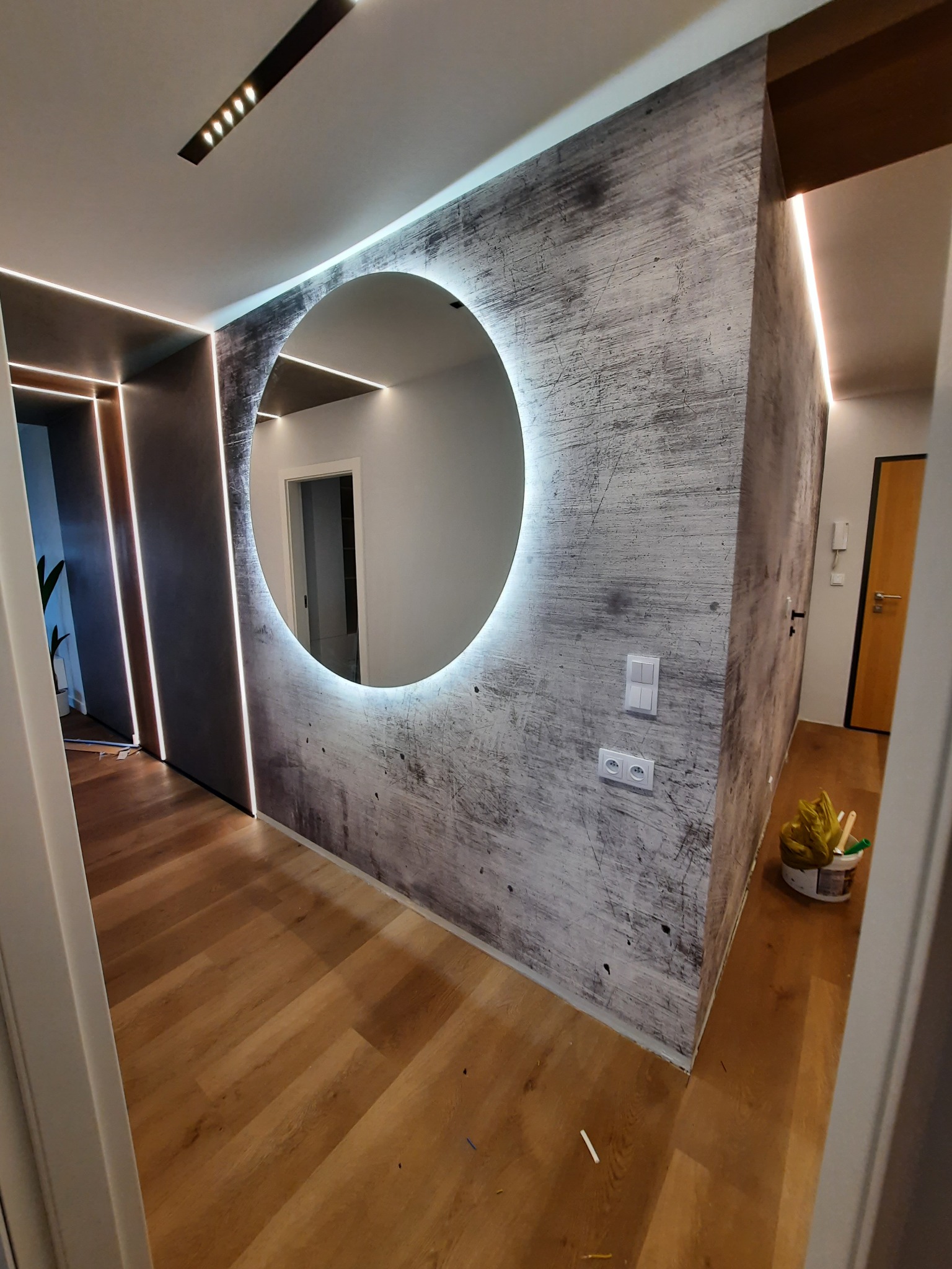 Nowoczesny przedpokój z okrągłym lustrem podświetlanym LED, ściany z efektem betonu, drewniana podłoga i oświetlenie liniowe LED wzdłuż ścian.