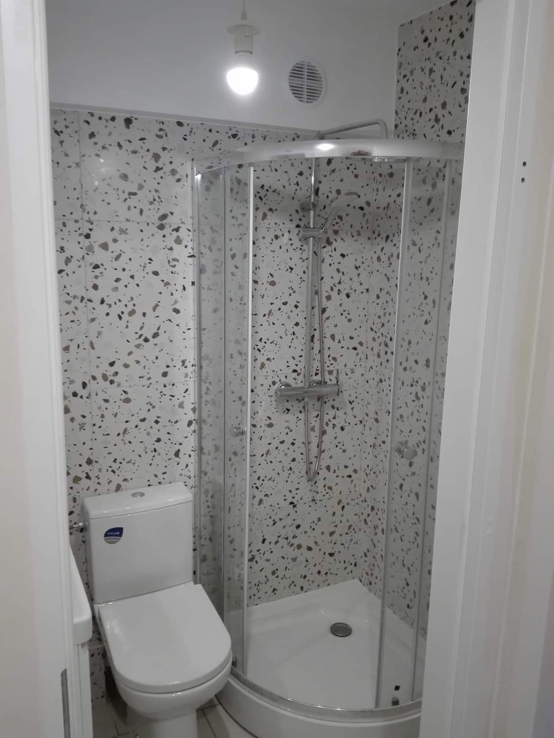 Nowoczesna łazienka z kabiną prysznicową narożną i toaletą, ściany wykończone płytkami w mozaikę z nieregularnymi plamkami, oświetlenie sufitowe punktowe.