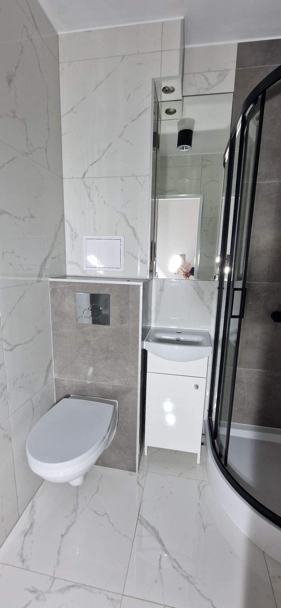 Nowoczesna łazienka po remoncie z marmurowymi płytkami, podwieszaną toaletą, umywalką z szafką i kabiną prysznicową z czarnymi ramami.