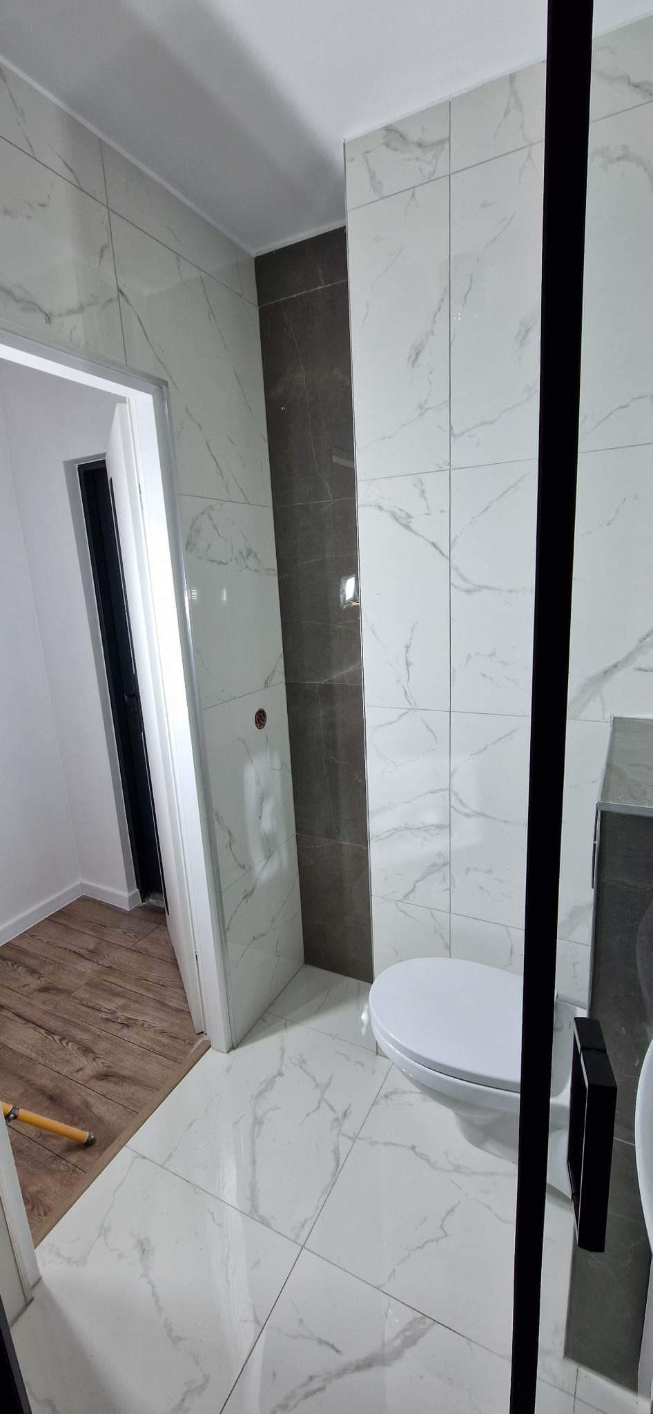 Wykończona łazienka z białymi i szarymi marmurkowymi płytkami, widoczne WC i fragment kabiny prysznicowej z czarnymi profilami oraz drewniana podłoga w sąsiednim pomieszczeniu.