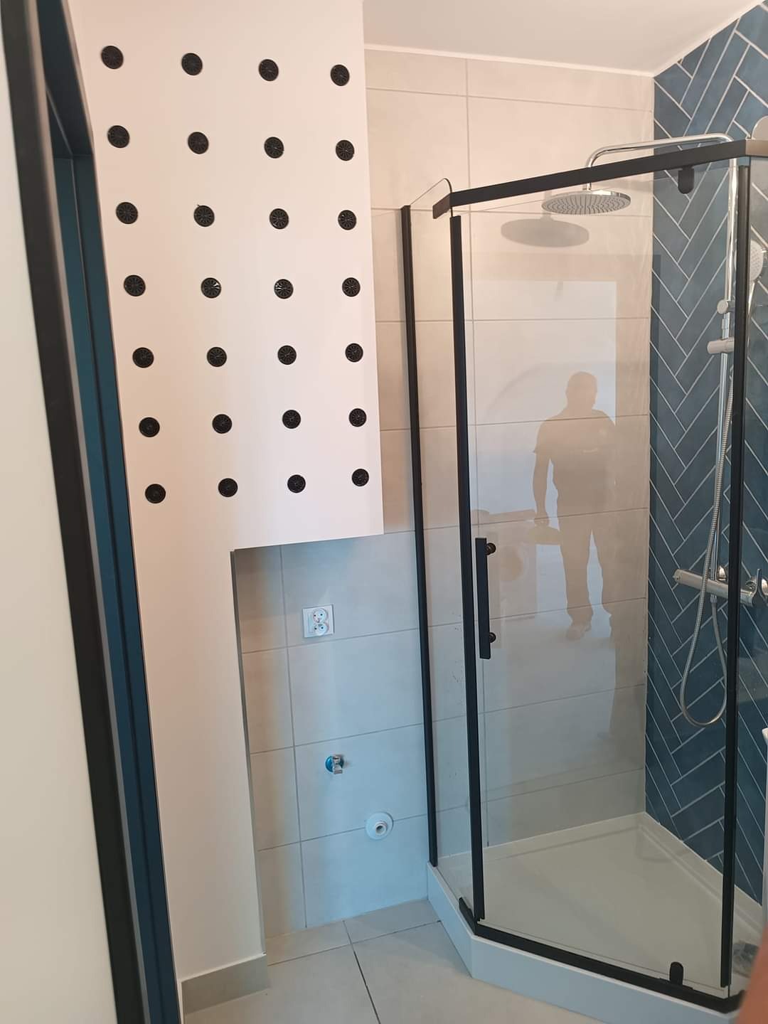 Nowoczesna kabina prysznicowa z czarnymi profilami, niebieska ściana w jodełkę, ściana z dekoracyjnymi czarnymi kropkami, widoczne przyłącza wodne i gniazdko elektryczne.