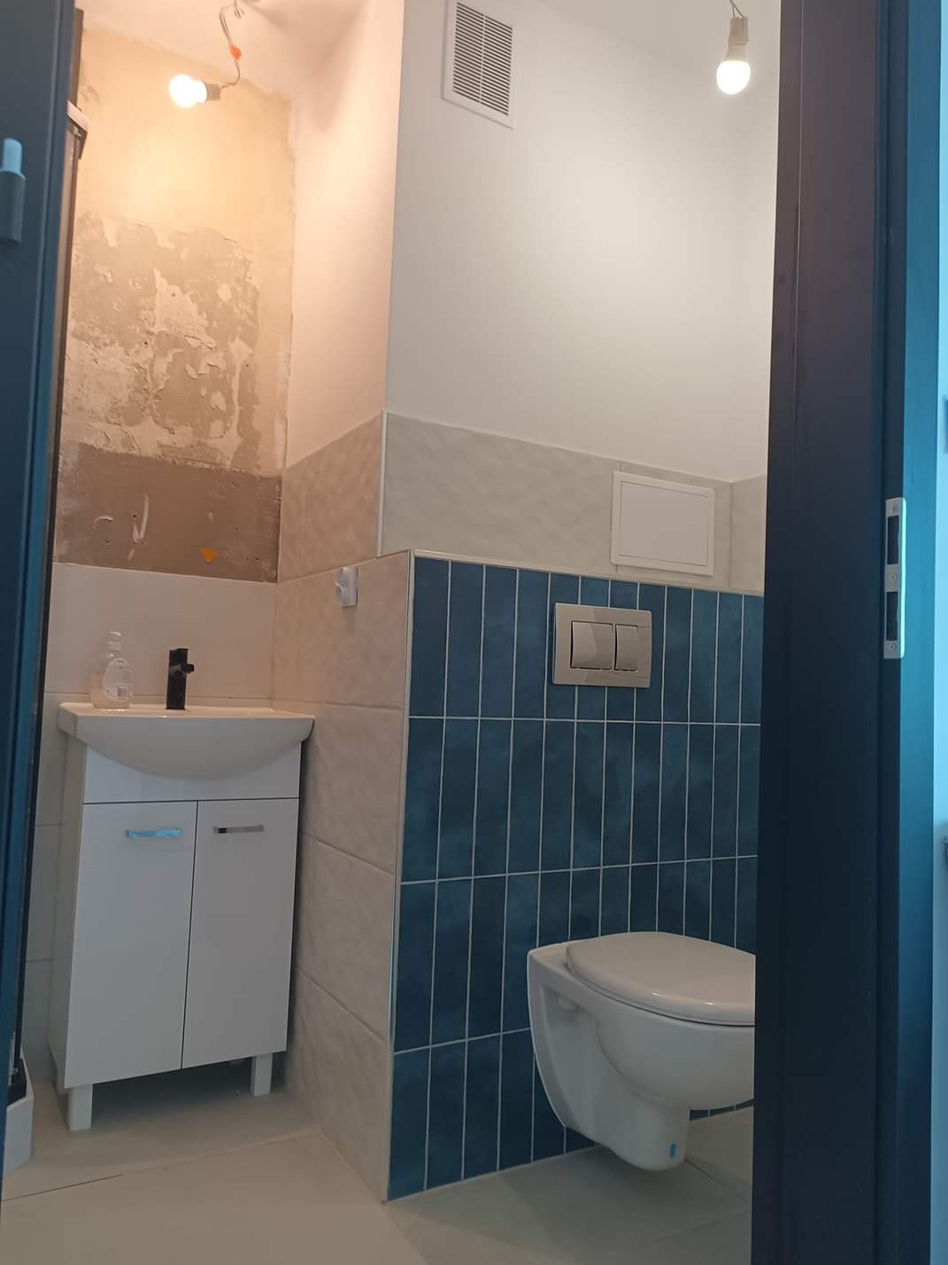 Niedokończona łazienka z białą szafką pod umywalką, wiszącą toaletą na tle niebieskich płytek i fragmentem ściany z odsłoniętym tynkiem, widok przez otwarte drzwi.