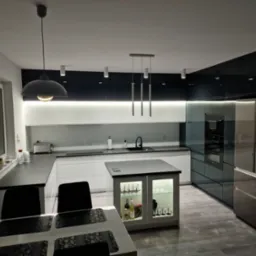 Nowoczesna kuchnia w odcieniach bieli i grafitu z wyspą, oświetleniem punktowym i wiszącą lampą nad stołem jadalnym. W tle widoczny sprzęt AGD i dekoracje okienne.