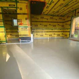 Świeżo wylana, gładka posadzka w pomieszczeniu z ocieplonymi ścianami i sufitem, widoczne okno balkonowe i urządzenie do rozprowadzania wylewki.