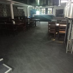 UkrPolWit - Wnętrze lokalu gastronomicznego z ciemnymi płytkami na podłodze, drewnianymi stolikami i krzesłami, wzorzystą tapetą na ścianie oraz barem z podświetleniem w tle.