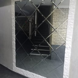 UkrPolWit - Dekoracyjne lustro złożone z romboidalnych elementów, obramowane imitacją białej cegły, odbijające wnętrze z schodami i ścianą z cegły.