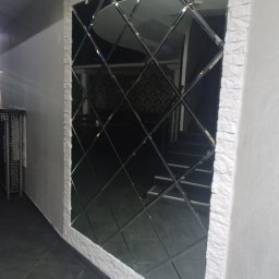 UkrPolWit - Ściana z dekoracyjnym lustrem w romby, obramowana imitacją kamienia, odbijająca schody i wzorzystą tapetę w tle.