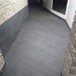 UkrPolWit - Wykończone wnętrze z szarymi płytkami podłogowymi ułożonymi diagonalnie, ściany częściowo wyłożone dekoracyjną cegłą i szarymi płytami, w tle widoczny mebel z greckim wzorem.