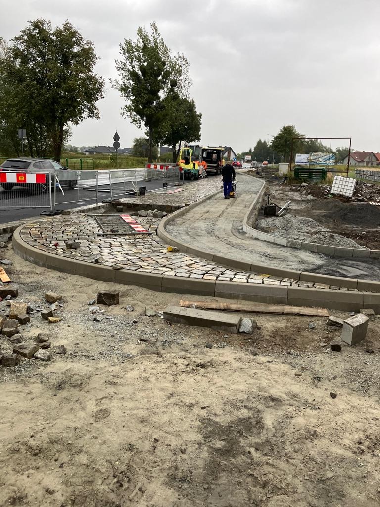Ułożona kostka brukowa w łuku z widocznym fragmentem drogi i pracownikiem zagęszczającym podłoże, maszyny budowlane w tle, pochmurne niebo.