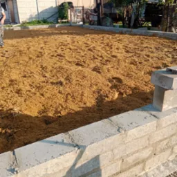 Wyrównany teren piaskiem wewnątrz częściowo wymurowanego ogrodzenia z betonowych bloczków, widoczne ślady stóp na piasku, w tle zabudowania gospodarcze.