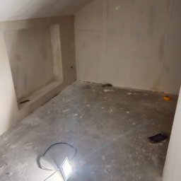 Dawid bud - Wnętrze pokoju na poddaszu w Wydrzynie w trakcie prac wykończeniowych, widoczna wnęka w ścianie i lampa robocza na podłodze.