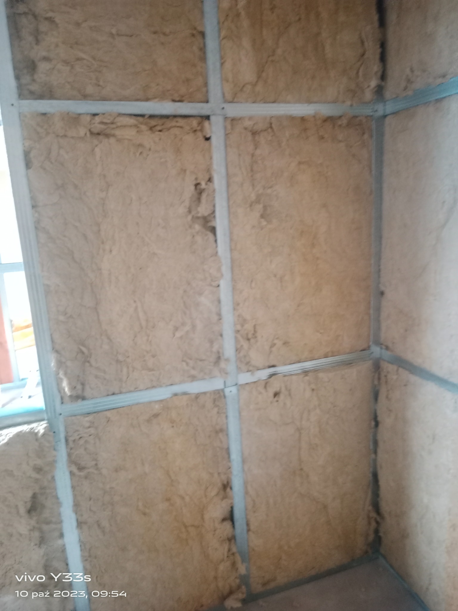 Ściana wewnętrzna z wełną mineralną, widoczne metalowe profile konstrukcyjne, ujęcie z dołu w słabym oświetleniu.