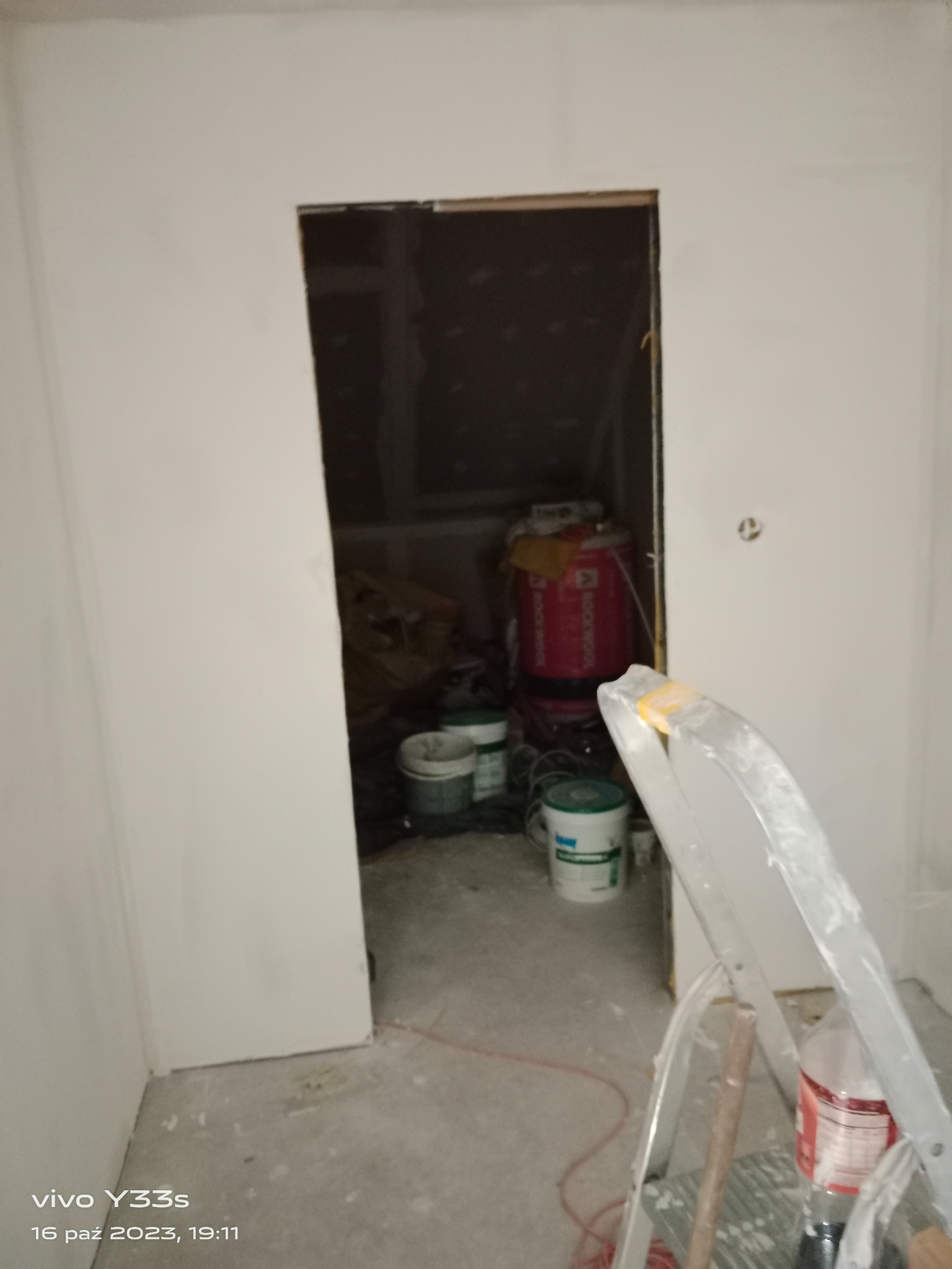 Wnętrze w trakcie prac wykończeniowych: białe ściany, otwór drzwiowy bez ościeżnicy, widoczne wiadro z farbą, puszka z wełną mineralną i drabina malarska.