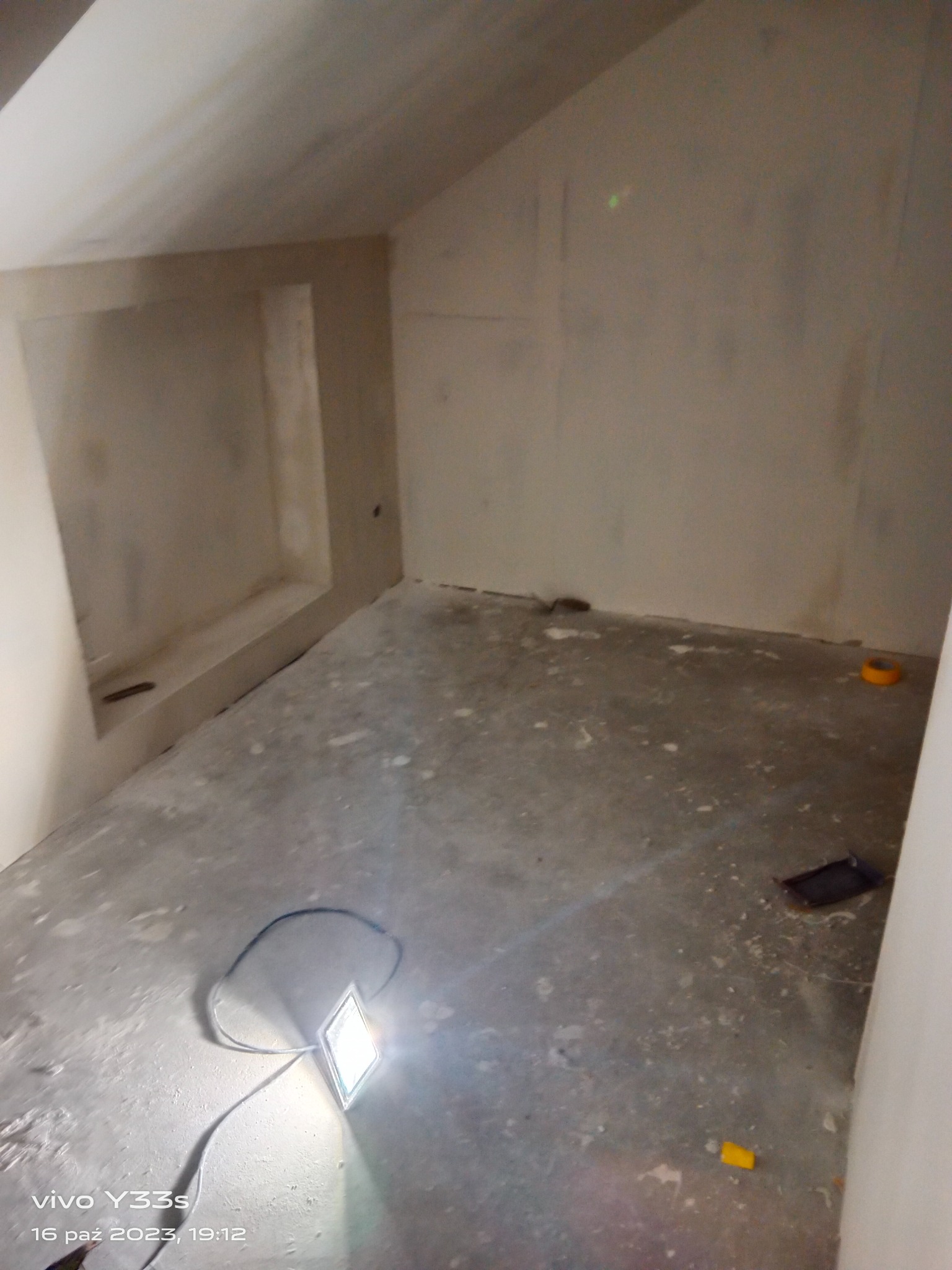 Wnętrze pokoju na poddaszu w Wydrzynie w trakcie prac wykończeniowych, widoczna wnęka w ścianie i lampa robocza na podłodze.