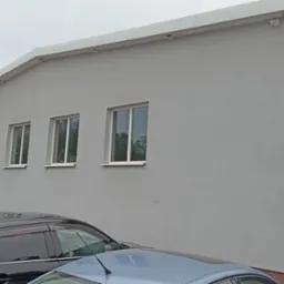 Jasnoszara elewacja budynku z czterema oknami w Końskich, widoczna z poziomu parkingu z zaparkowanymi samochodami. Na rogu budynku rynna i kamera monitoringu.