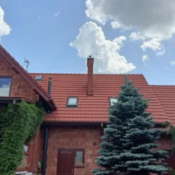 Dwupiętrowy dom z czerwonej cegły z czerwoną dachówką, zarośnięty zielenią po lewej stronie, na tle błękitnego nieba z białymi chmurami.