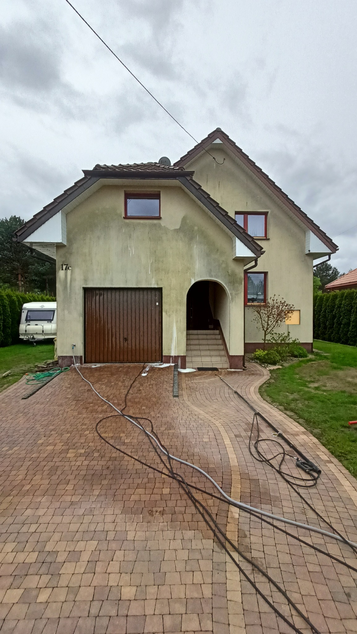 Dom jednorodzinny z garażem, elewacją w odcieniach beżu i brązu, częściowo umyta kostka brukowa na podjeździe, widoczne węże ciśnieniowe i ślady mycia na elewacji i podjeździe.