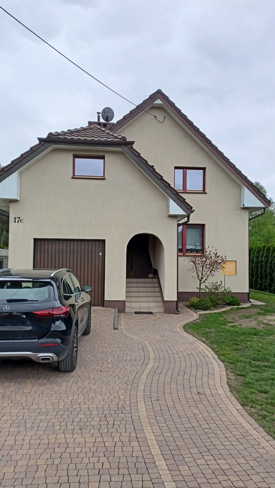Dom jednorodzinny z beżową elewacją, brązowymi ramami okiennymi i dachem z brązowej dachówki. Przed domem ułożona kostka brukowa w odcieniach brązu i beżu, zaparkowany czarny samochód osobowy.