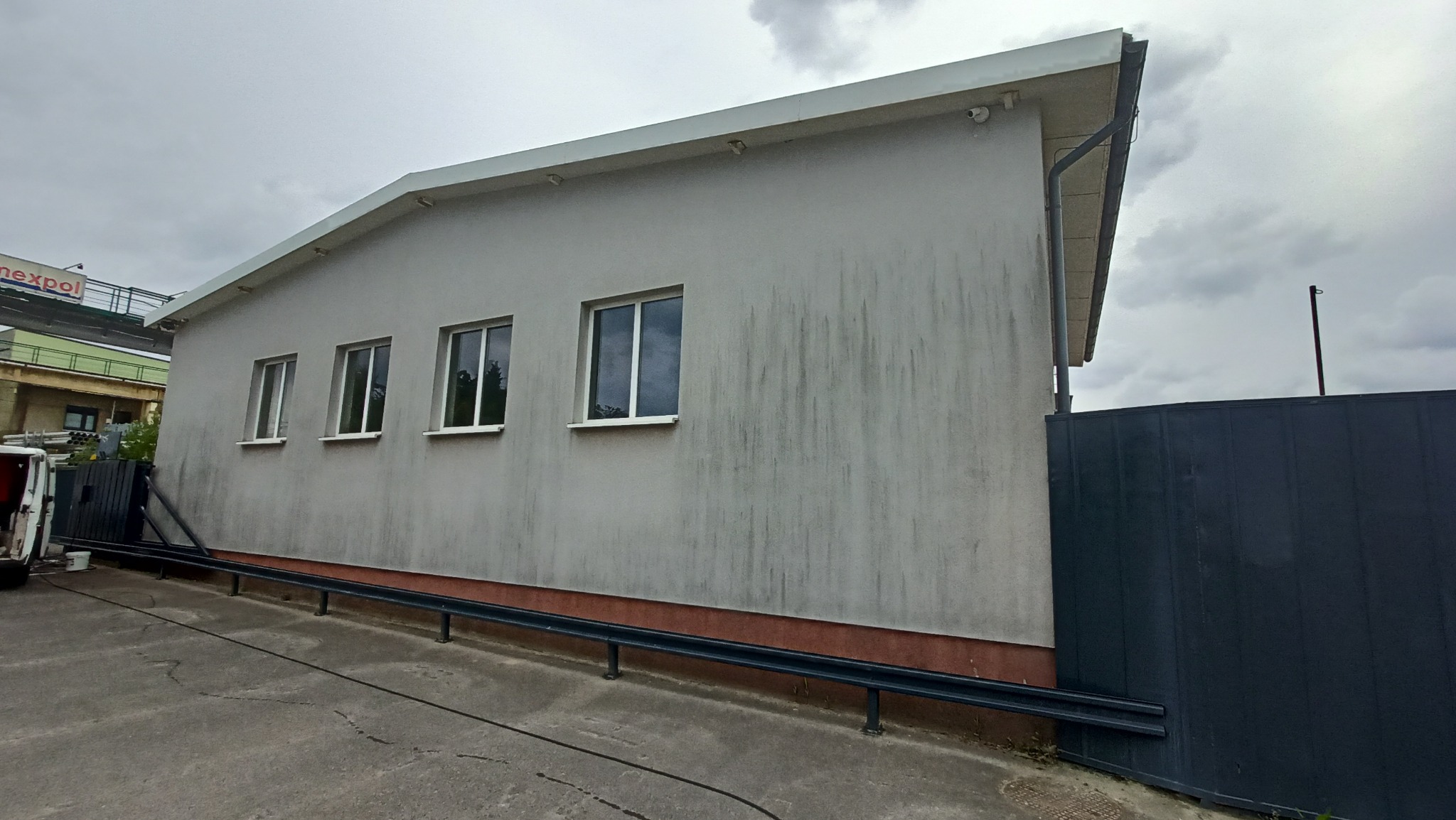 Szara, zabrudzona elewacja budynku z trzema oknami, widoczna z poziomu chodnika, z ciemnoszarą bramą po prawej stronie i fragmentem samochodu dostawczego po lewej.