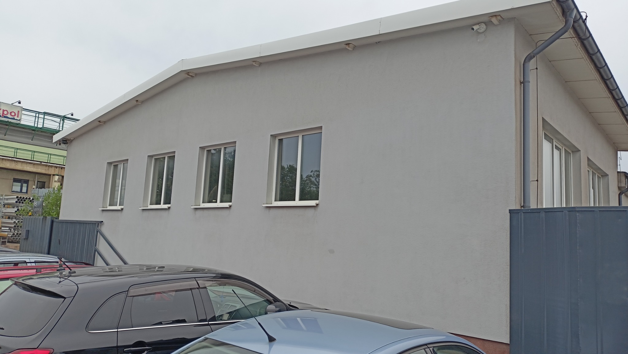 Jasnoszara elewacja budynku z czterema oknami w Końskich, widoczna z poziomu parkingu z zaparkowanymi samochodami. Na rogu budynku rynna i kamera monitoringu.