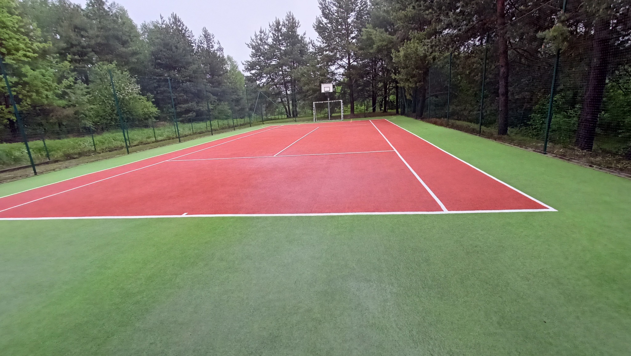 Czerwono-zielone boisko sportowe z bramką do piłki nożnej i siatką ochronną, otoczone drzewami.