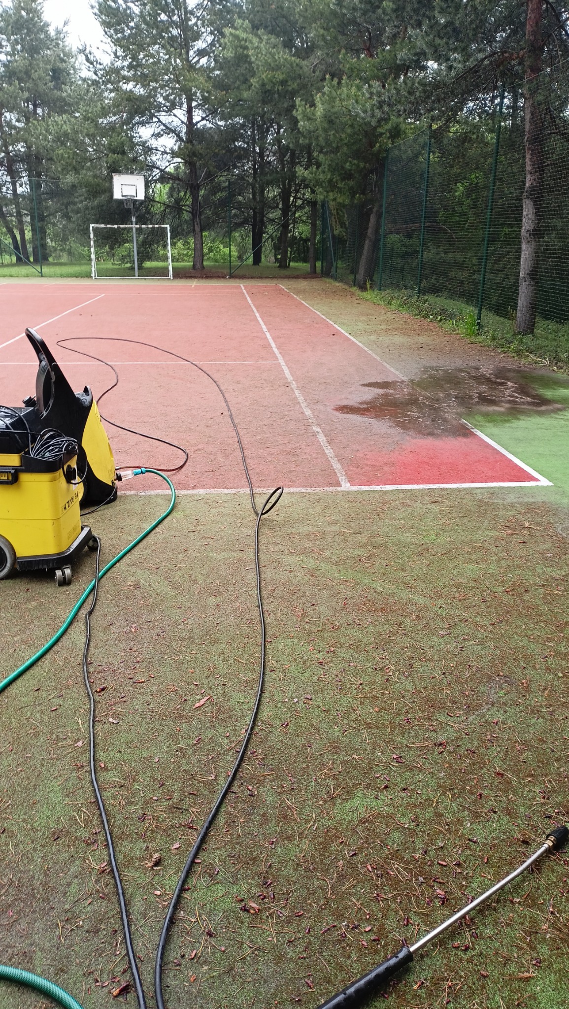 Żółta myjka ciśnieniowa z wężami na boisku sportowym z czerwoną nawierzchnią i zielonym obrzeżem, widoczne ślady mycia oraz bramka i kosz do gry w tle.
