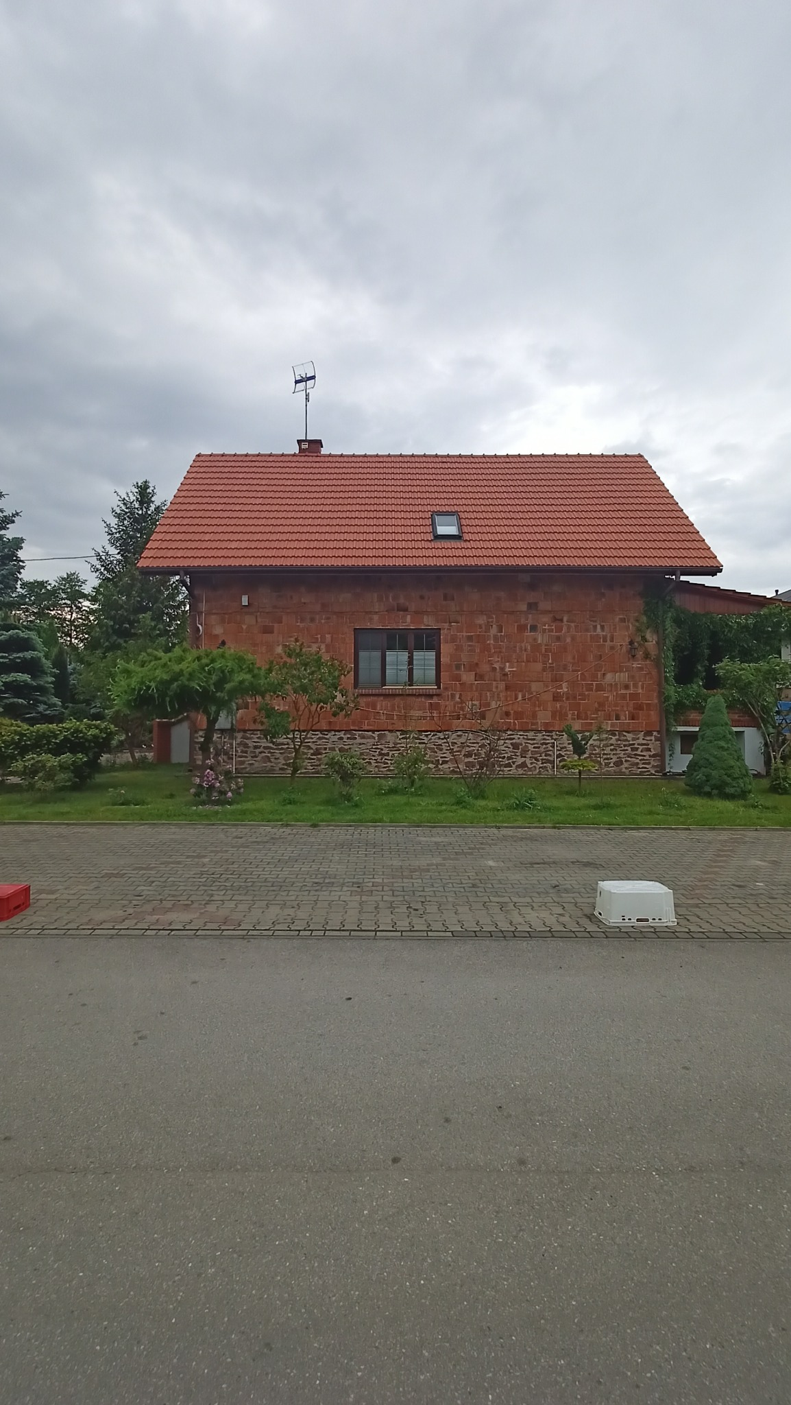 Dom jednorodzinny z czerwonej cegły z czerwoną dachówką, widok z poziomu ulicy w pochmurny dzień. Antena telewizyjna na dachu.
