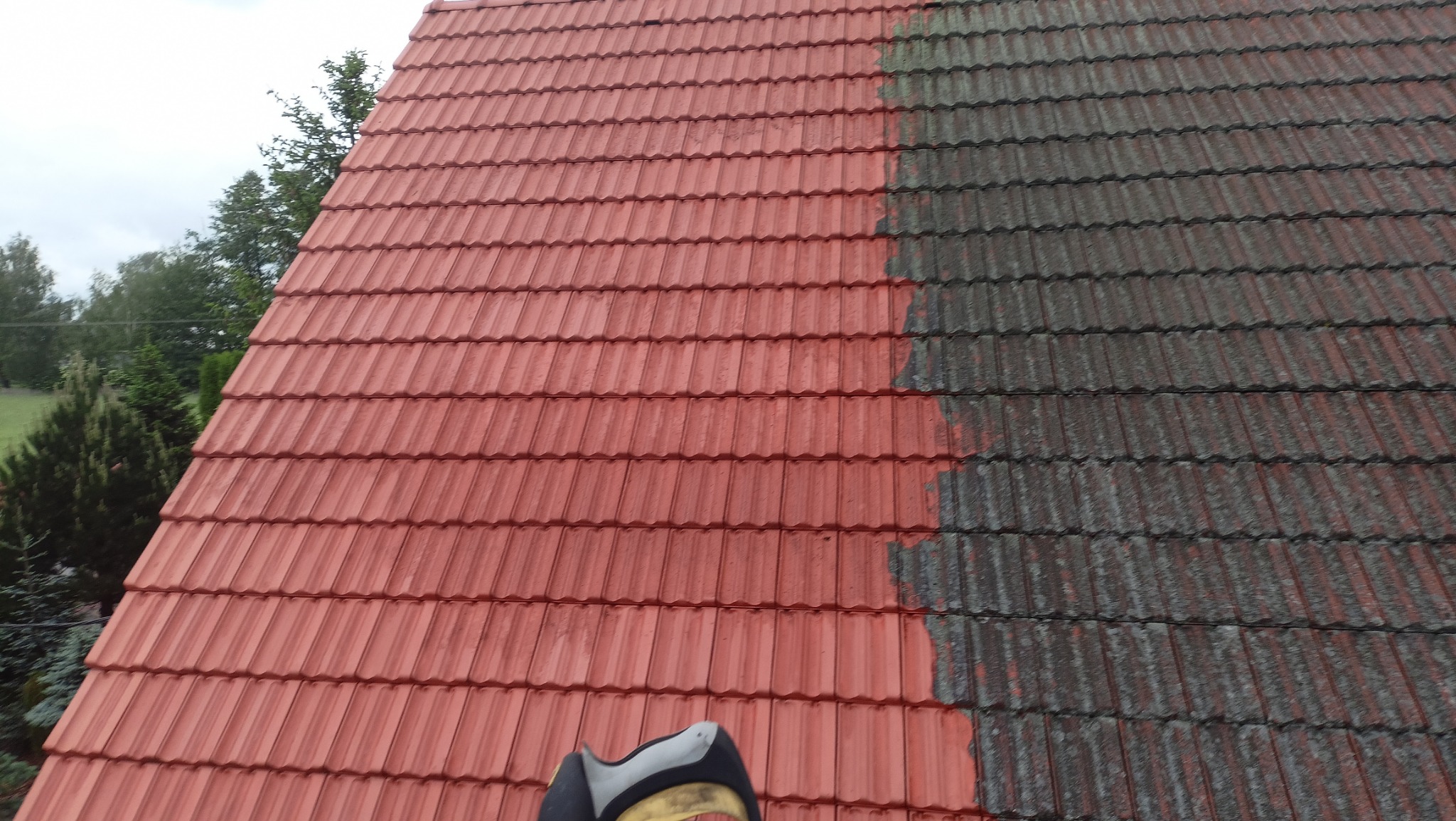 Częściowo oczyszczony dach z czerwonej dachówki, widoczna wyraźna różnica między umytą a zabrudzoną powierzchnią, fragment narzędzia w dolnej części kadru.