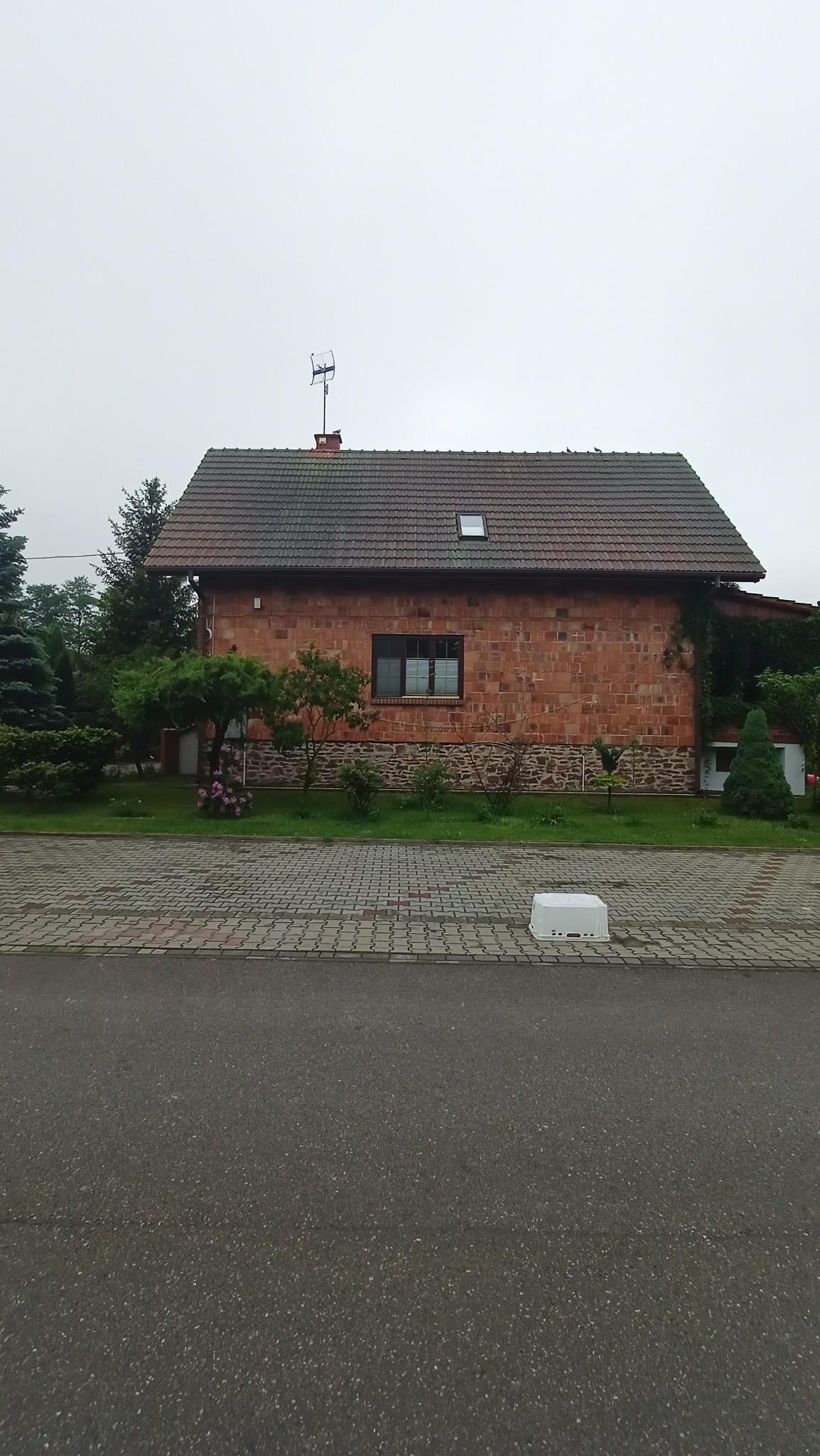 Dwukondygnacyjny dom z czerwonej cegły z ciemną dachówką i anteną na dachu, widziany z poziomu ulicy, z kostką brukową i zielenią w tle.