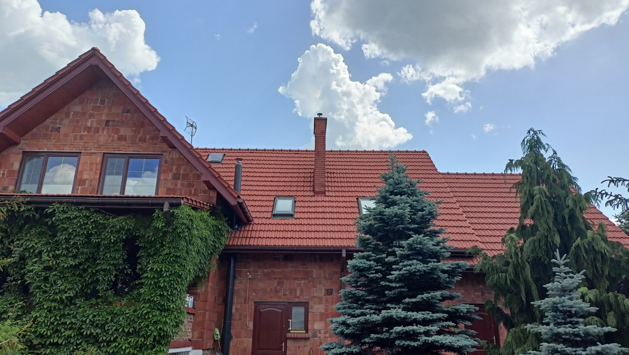 Dwupiętrowy dom z czerwonej cegły z czerwoną dachówką, zarośnięty zielenią po lewej stronie, na tle błękitnego nieba z białymi chmurami.