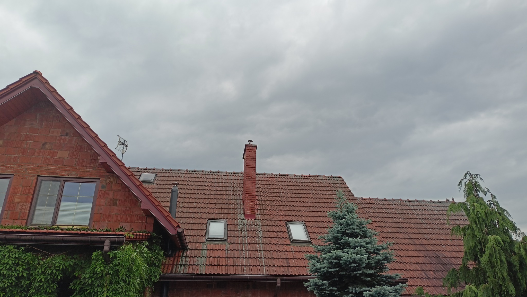 Ceglany dom z czerwonym dachem, kominem i anteną na tle pochmurnego nieba. Widoczne okna dachowe i fragmenty roślinności.