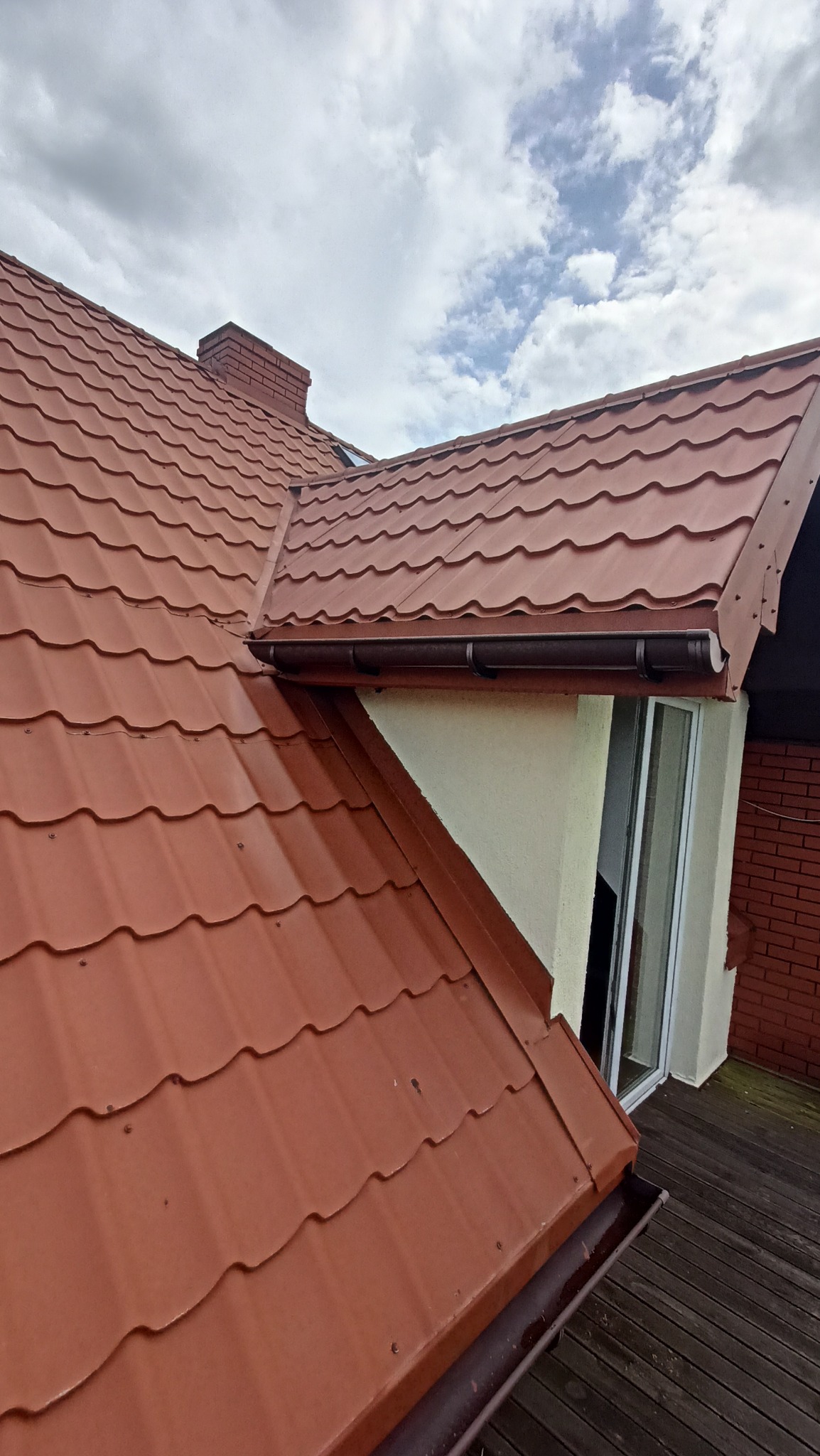Czerwony dach z blachy trapezowej, widoczny komin i rynna, na tle pochmurnego nieba. Dach z widocznymi śladami po deszczu.