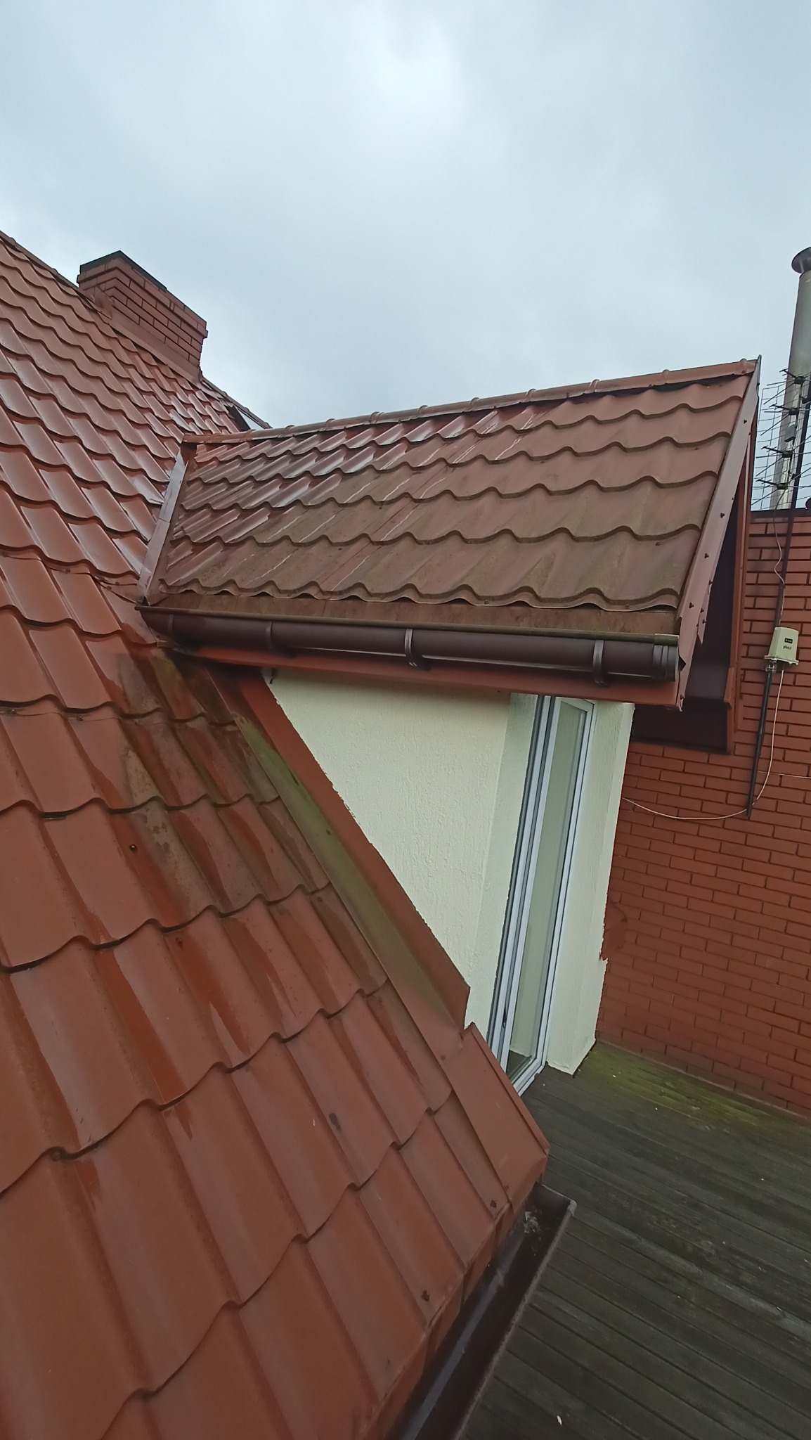 Czerwona dachówka na mokrym dachu, widoczny komin, rynna i fragment ściany budynku z oknem, ujęcie z góry po deszczu.