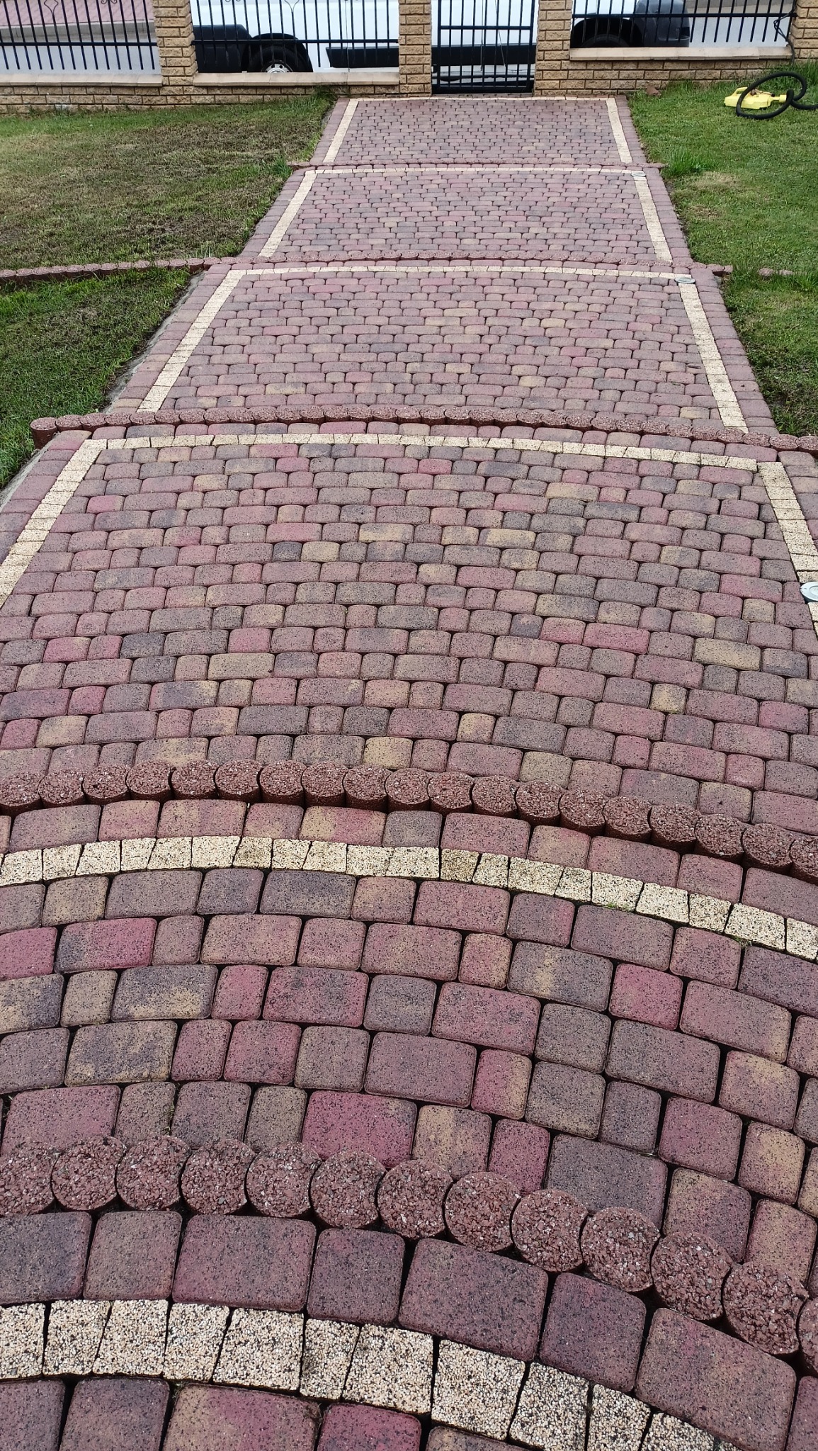 Ułożona z czerwono-brązowej kostki brukowej ścieżka w ogrodzie, z obrzeżami z okrągłych elementów i jasnych kostek, prowadząca do furtki w metalowym ogrodzeniu.