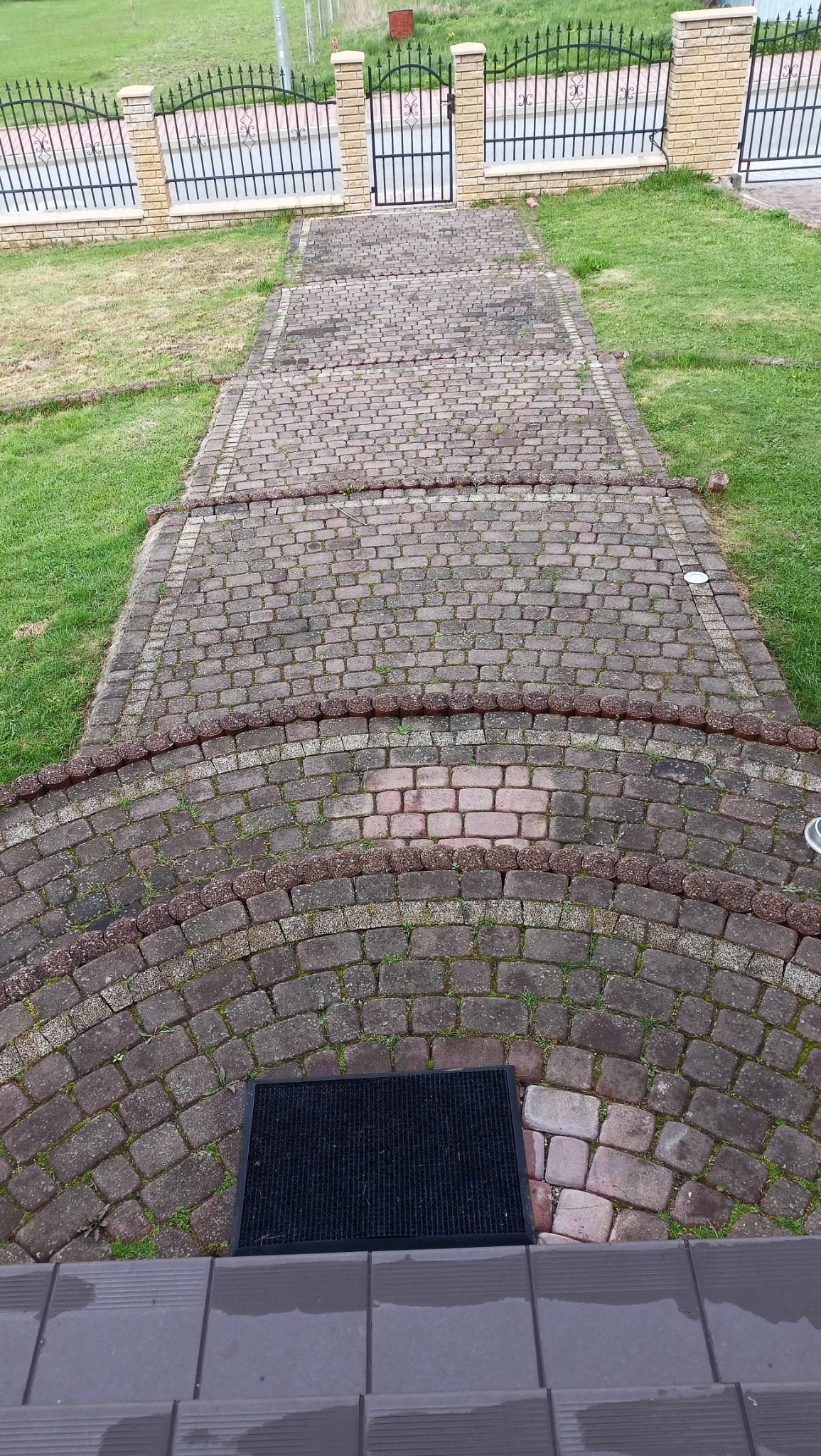 Ceglana ścieżka z czarną wycieraczką prowadząca do bramy z metalowymi przęsłami, otoczona trawnikiem z widocznymi śladami mchu i zabrudzeń.