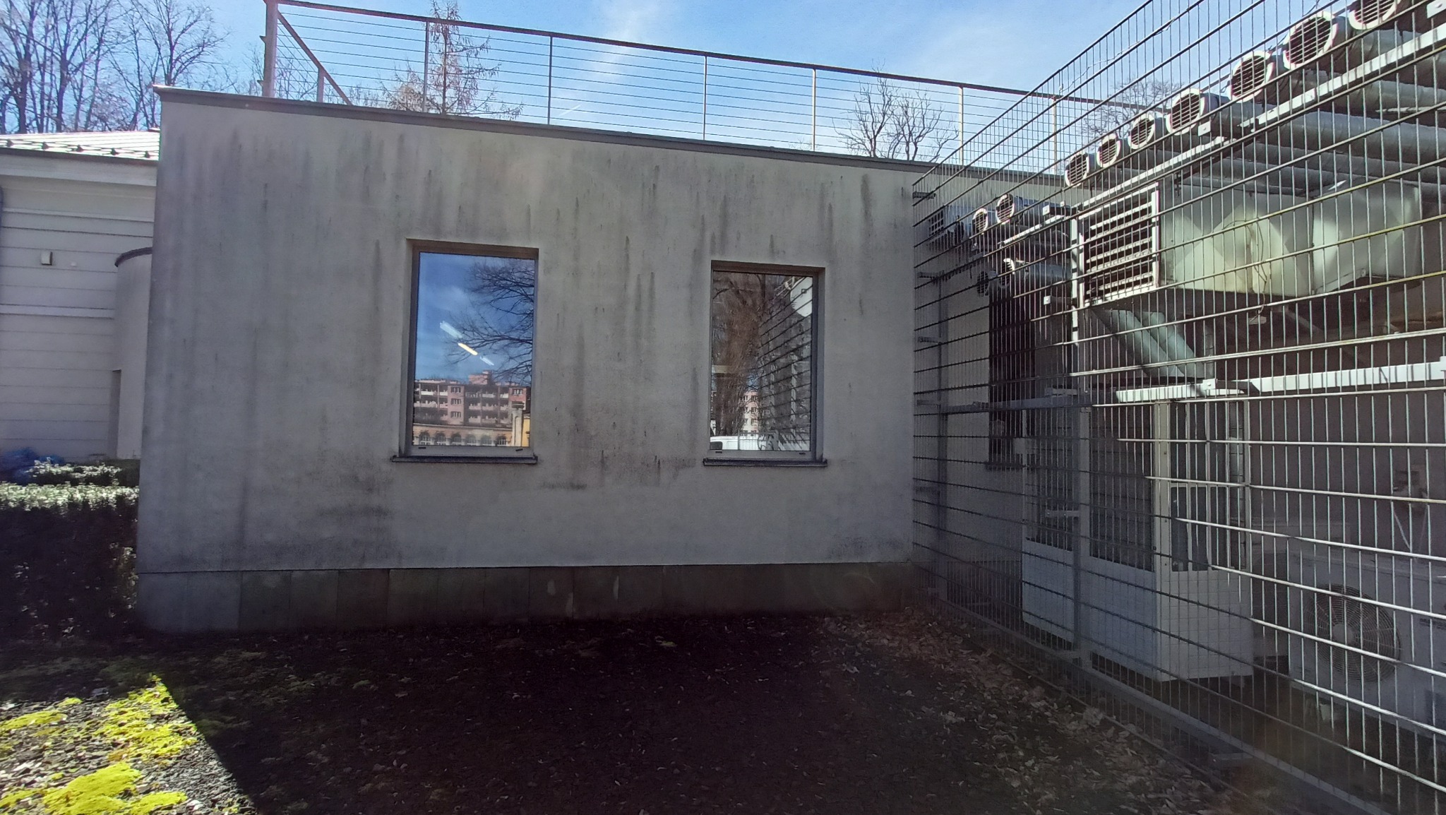 Szara, zabrudzona elewacja budynku z dwoma oknami, w których odbija się inny budynek. Po prawej stronie widoczna metalowa konstrukcja z urządzeniami wentylacyjnymi.