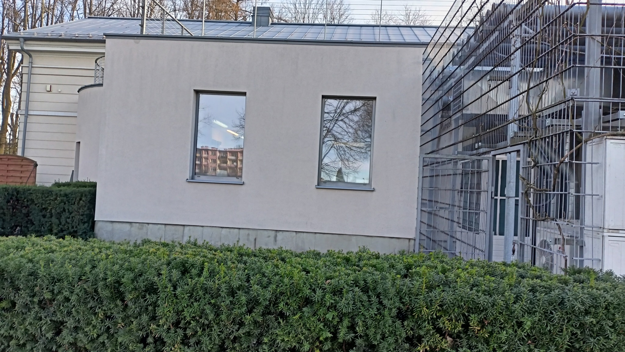 Jasnoszara elewacja budynku z dwoma oknami, w których odbija się budynek mieszkalny, z widocznym fragmentem metalowej konstrukcji po prawej stronie i niskim żywopłotem na pierwszym planie.