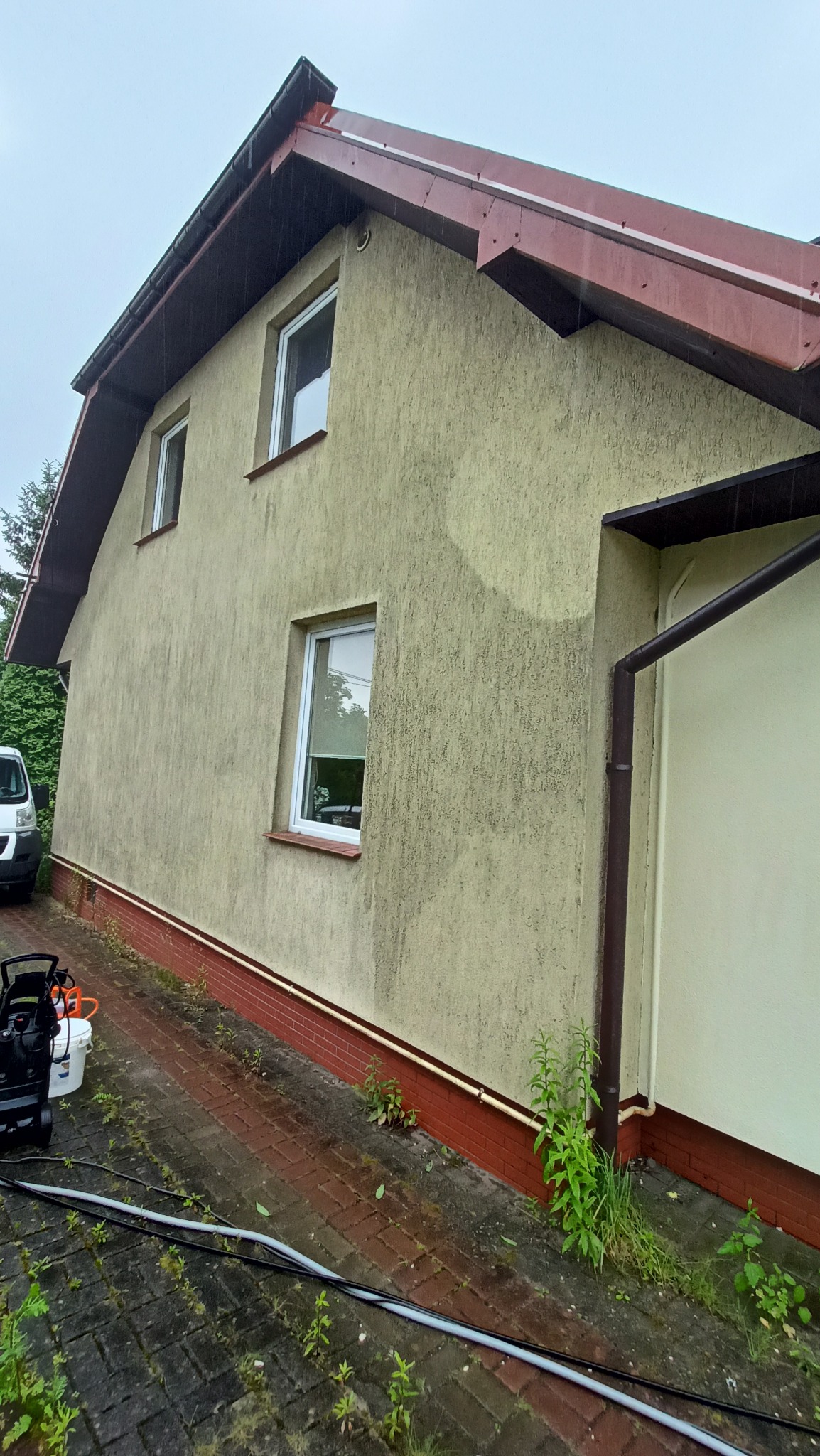 Fragment elewacji domu z widocznymi śladami po czyszczeniu, z myjką ciśnieniową i wiadrem na pierwszym planie, kostka brukowa częściowo porośnięta zielenią.