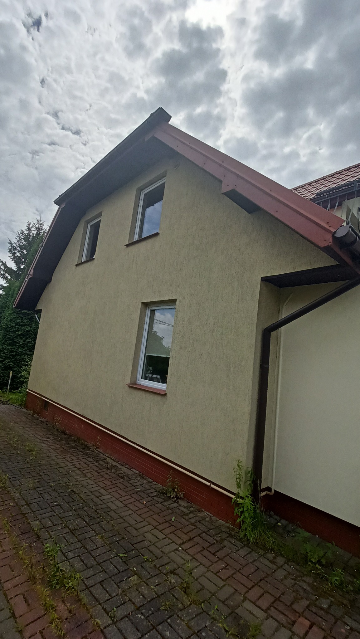 Boczny widok domu z elewacją w odcieniach beżu, widoczna kostka brukowa z chwastami u podstawy ściany. Dom posiada trzy okna z białymi ramami oraz brązowy dach i rynny. Niebo jest pochmurne.
